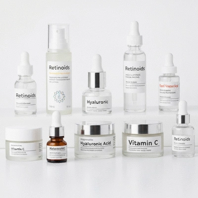 Close-up of various small bottles and jars of skincare products with ingredients like retinoids, hyaluronic acid, and vitamin C clearly labeled on a clean, bright background.